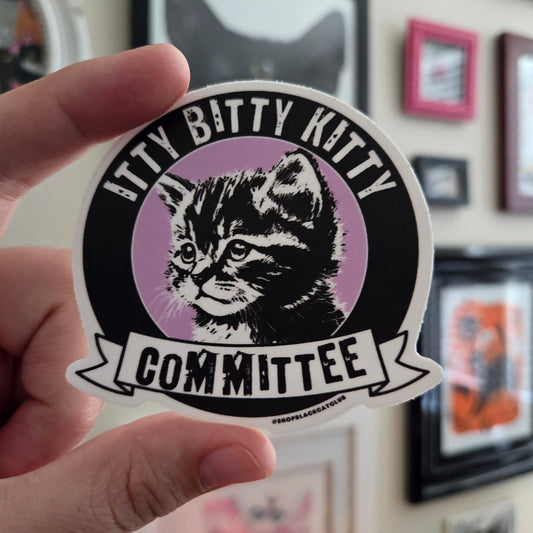 A round sticker with a black and purple design featuring an image of a cat and the text 'ITTY BITTY KITTY COMMITTEE'. The sticker is held by a hand against a background of framed artwork and decoration.
