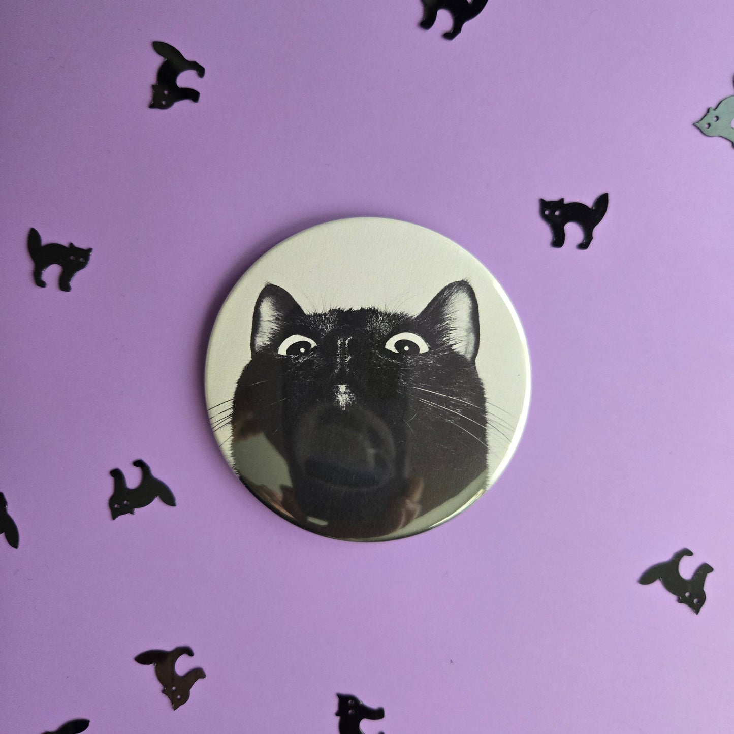 one circular magnets with a silly black cat photo, displayed on a purple background with small black cat confetti.