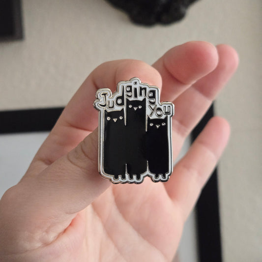 A hand holding up a black and white enamel pin reading Judging You with an illustration of 3 cats
