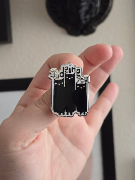 A hand holding up a black and white enamel pin reading Judging You with an illustration of 3 cats