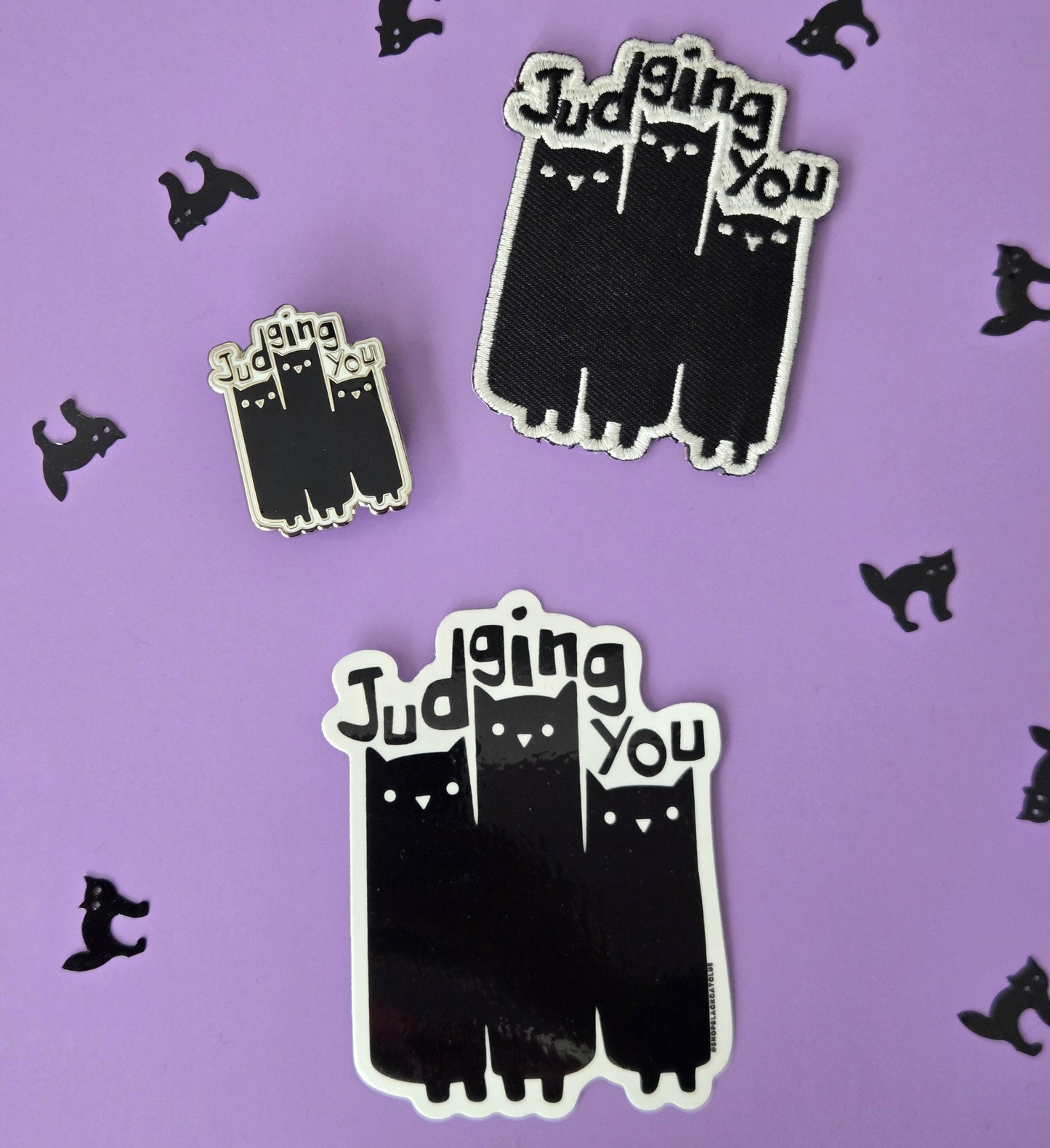 Black and white enamel pin, sticker, and patch reading Judging You with an illustration of 3 cats, displayed on a purple background with small black cat confetti.