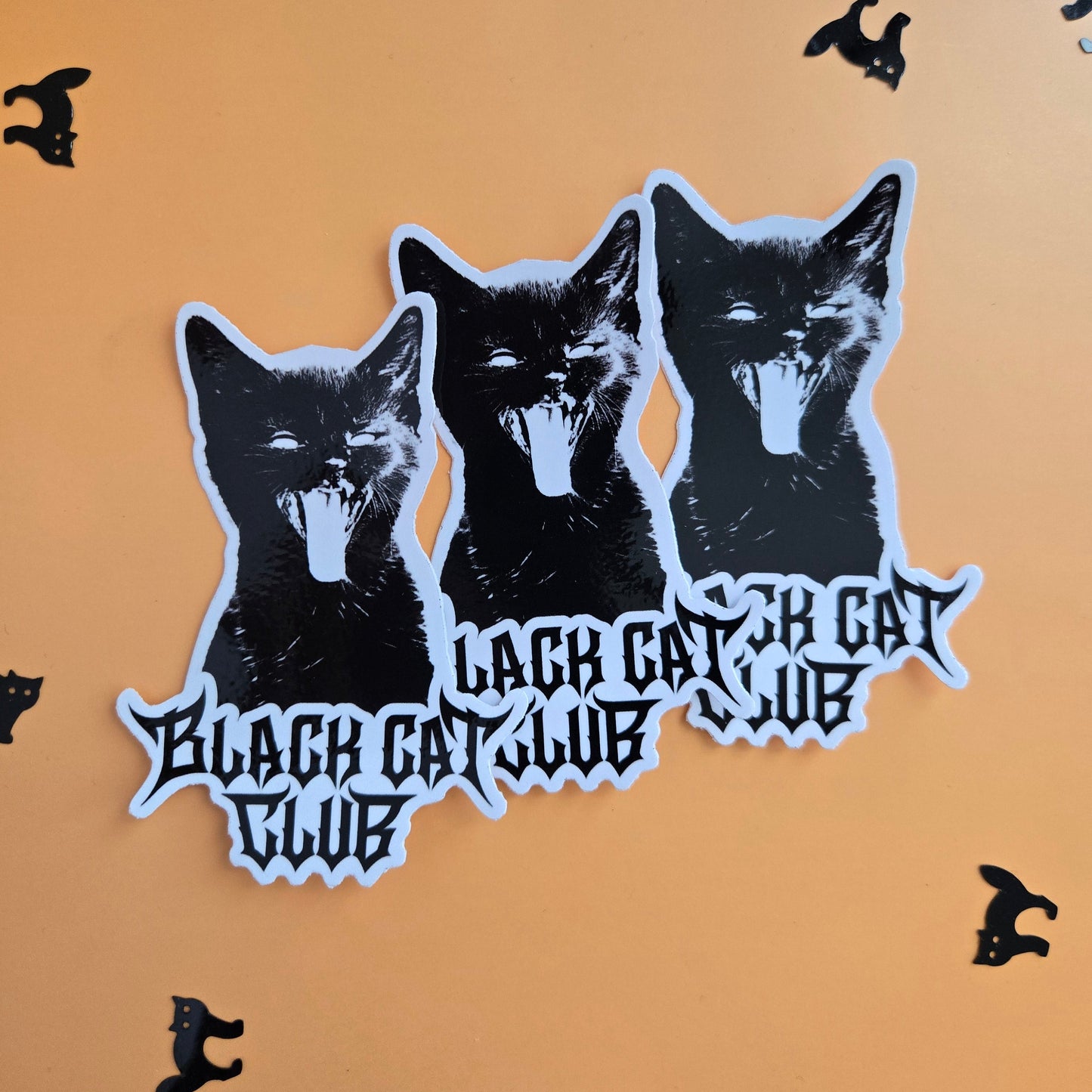 Three stacked black and white stickers featuring a stylized cat head and gothic text, displayed on a warm orange background with small black cat silhouettes scattered around.