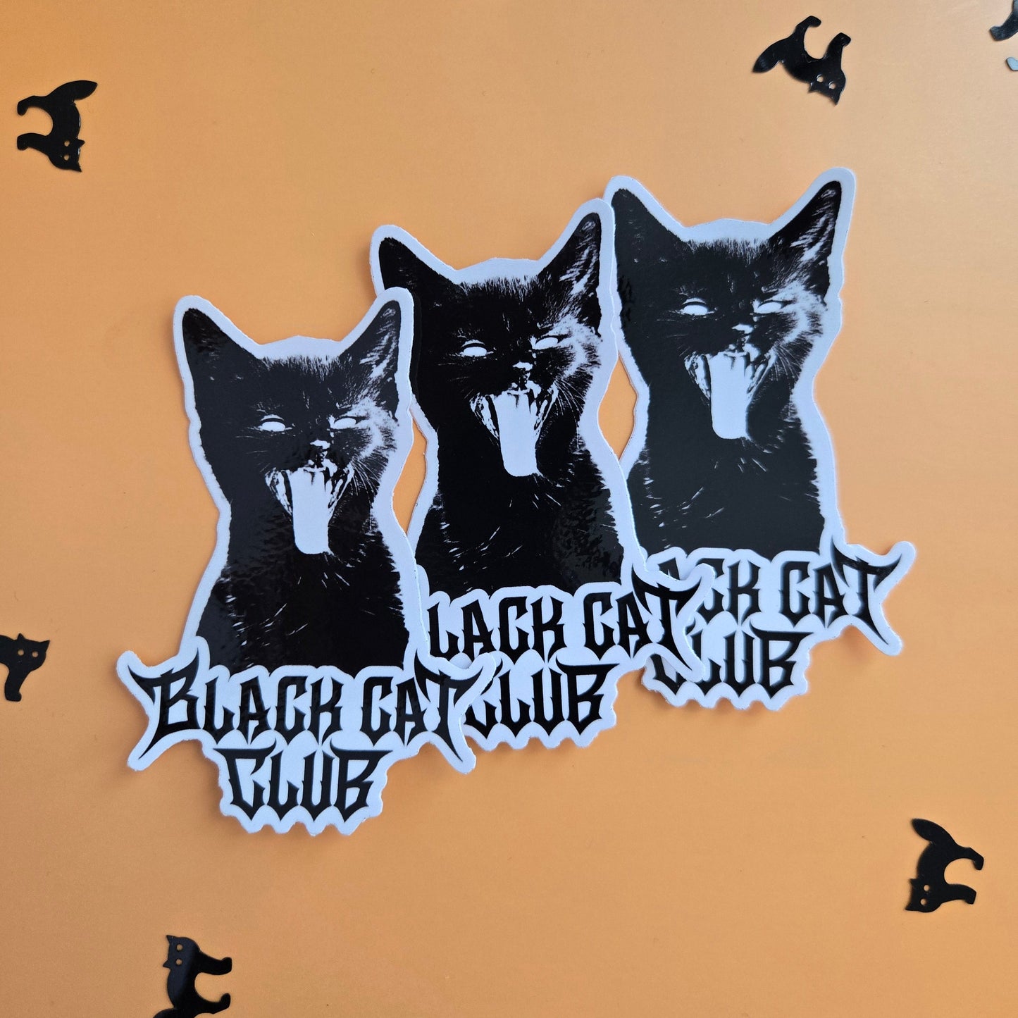 A die-cut sticker featuring three black cat heads with white eyes and gothic text reading 'Black Cat Club' on a peach background, surrounded by small black cat silhouettes.