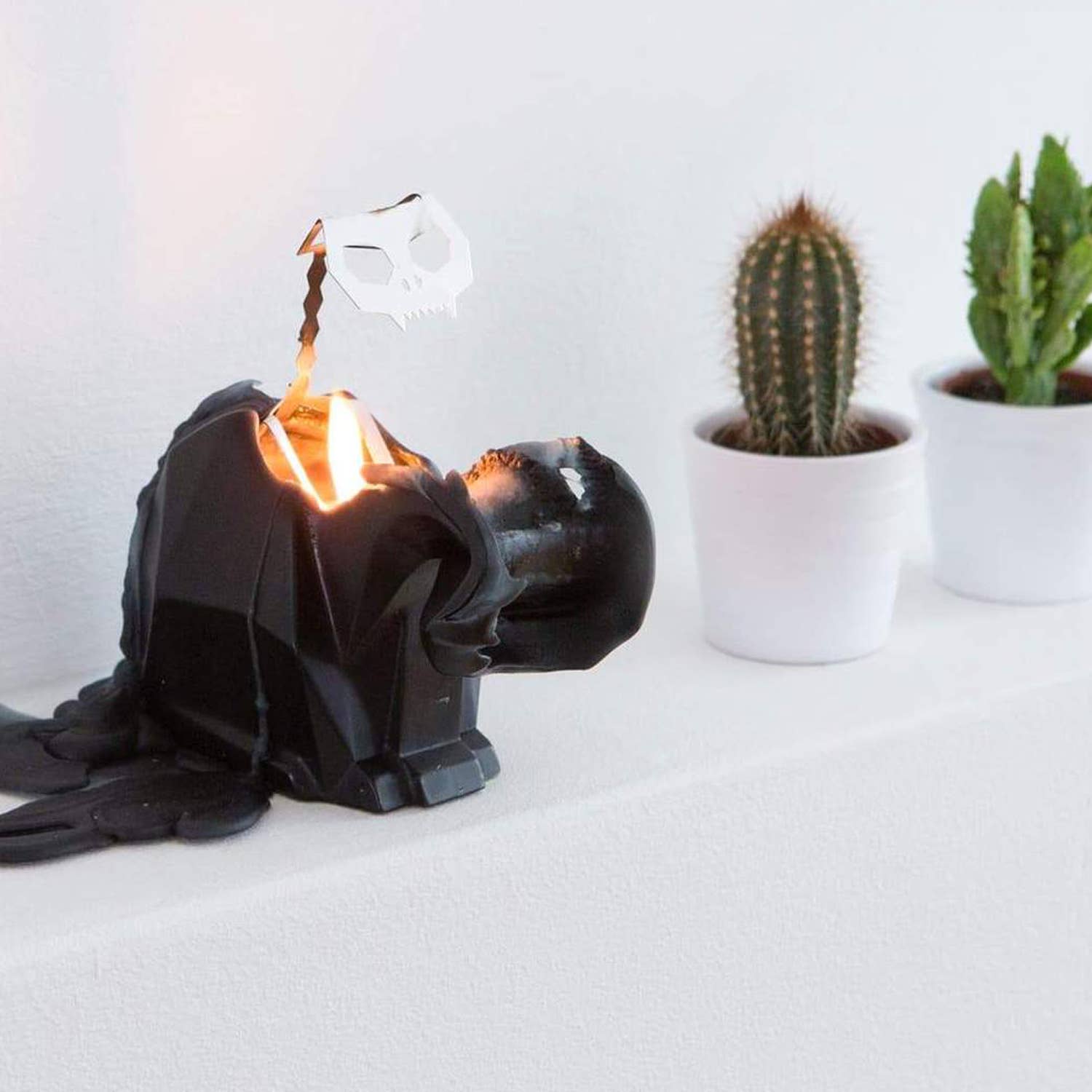 A black cat-shaped candle with a metallic skeleton design inside, partially melted to reveal the skeleton on a white background with a cactus in a pot