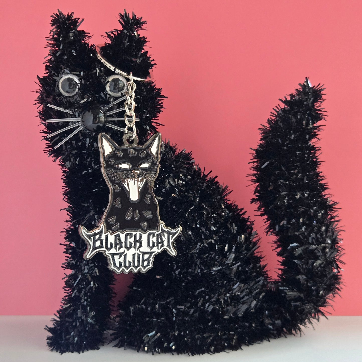 Black tinsel cat-shaped ornament with a black cat shaped keychain hanging off it's ear on a pink background