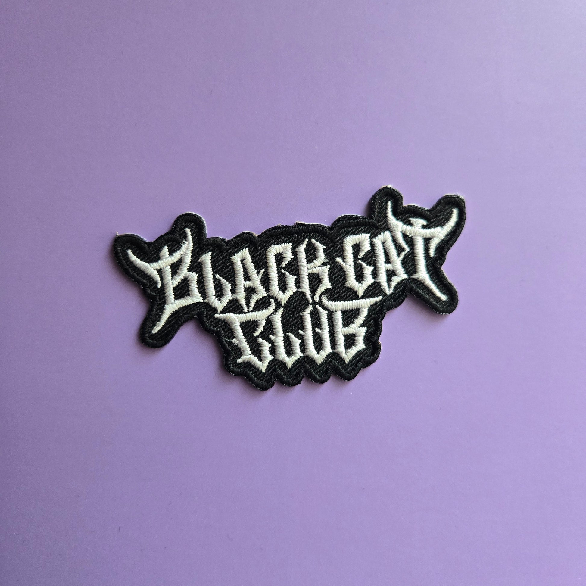 Black and white embroidered patch of the Black Cat Club logo on a purple background
