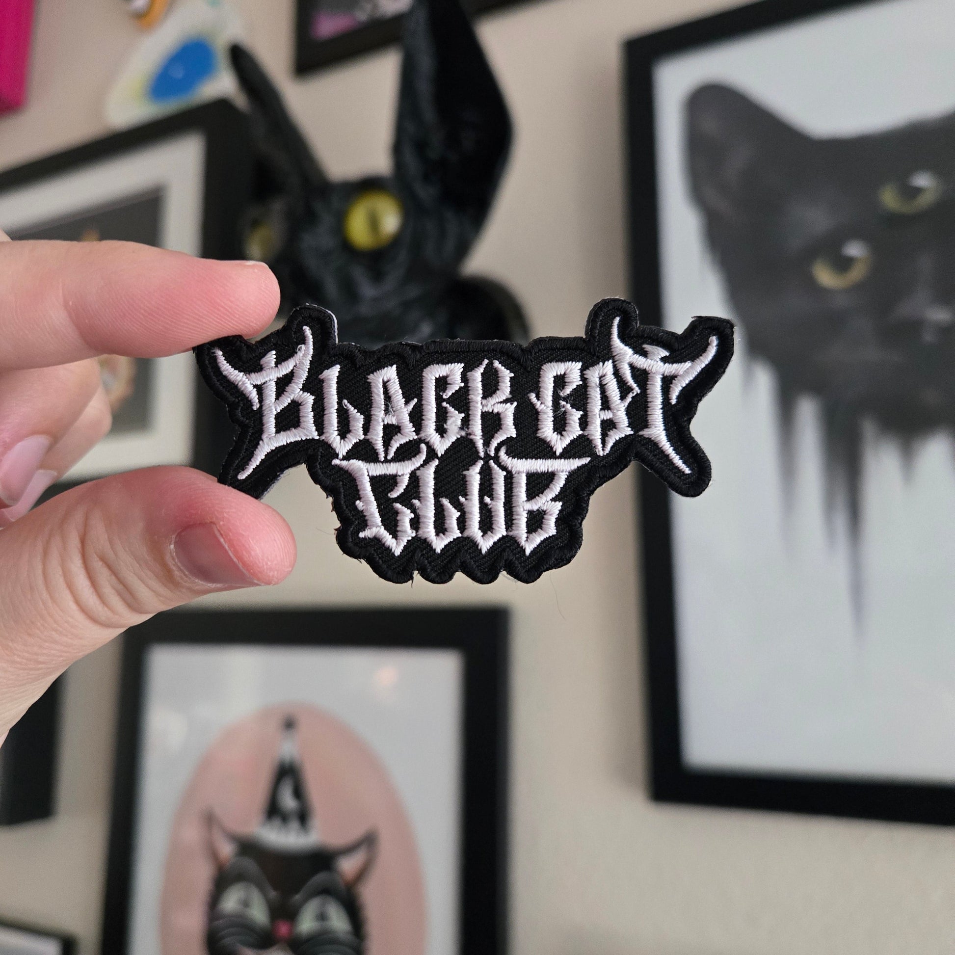 Hand holding a black and white cat-shaped patch with text against a wall with framed pictures.