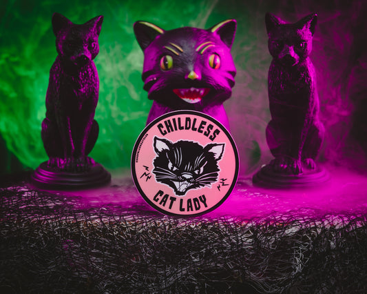 A round pink sticker with the text 'Childless Cat Lady' on it, depicted in a stylized font with black cat illustration, on a background featuring two cat figures and pink lighting.