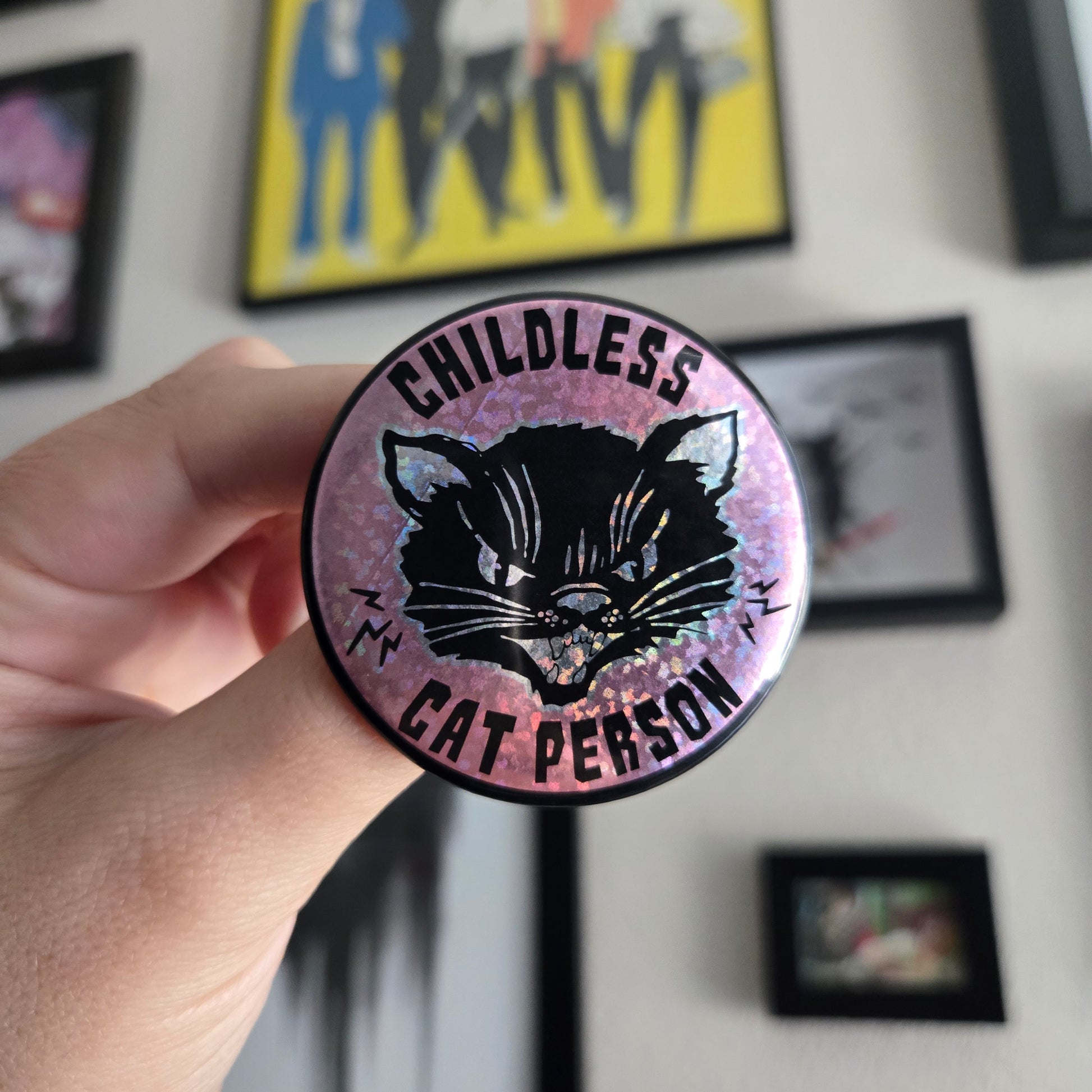 A hand holding a 2.25 inch round button with text 'Childless Cat Person' on a pink sparkle background, featuring a black cat.