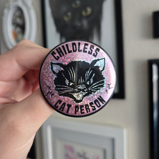 A hand holding a 2.25 inch round button with text 'Childless Cat Person' on a pink sparkle background, featuring a black cat.