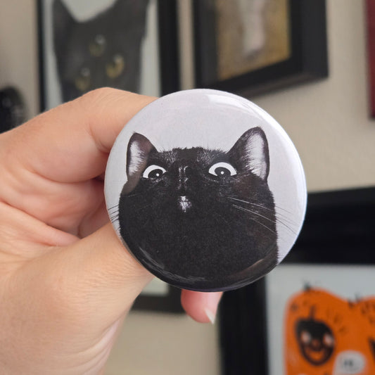 Button badge with a black cat illustration held by a hand in front of a blurred indoor background.