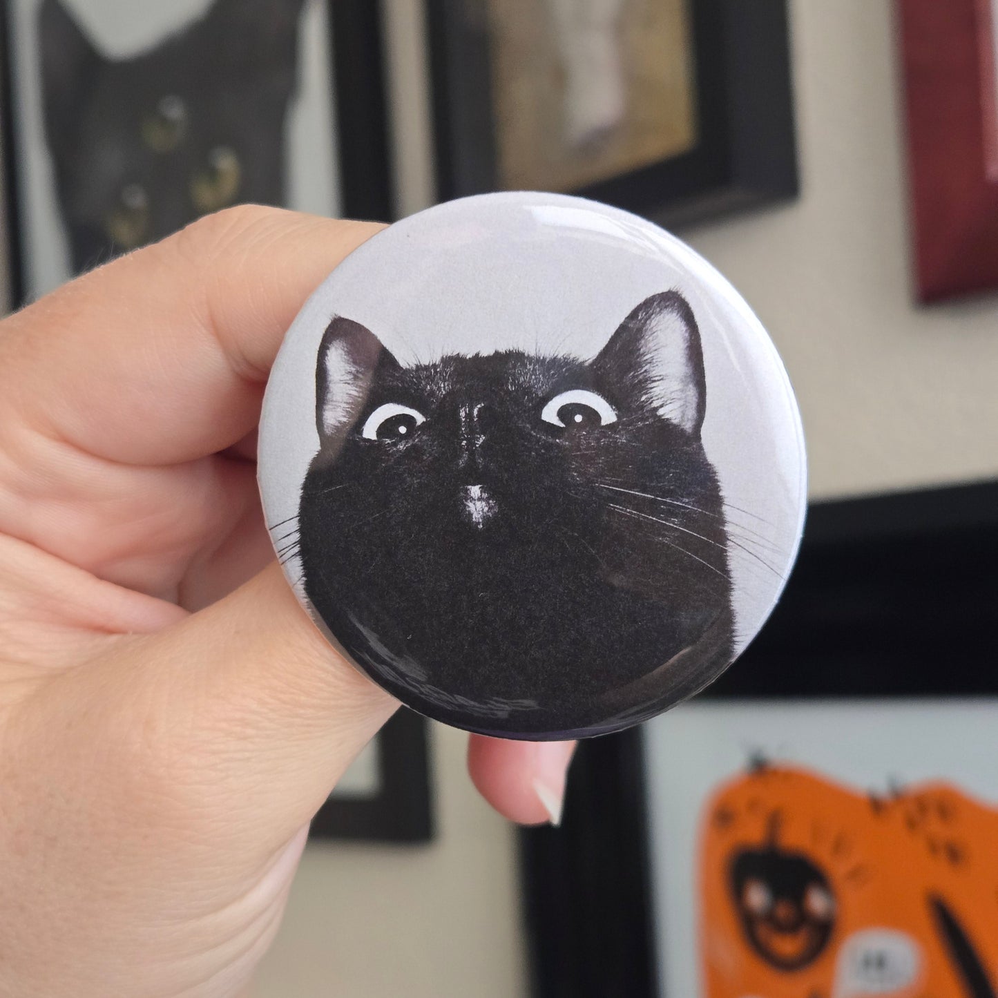 Round button badge with a black cat illustration held by a hand, with framed pictures in the background.