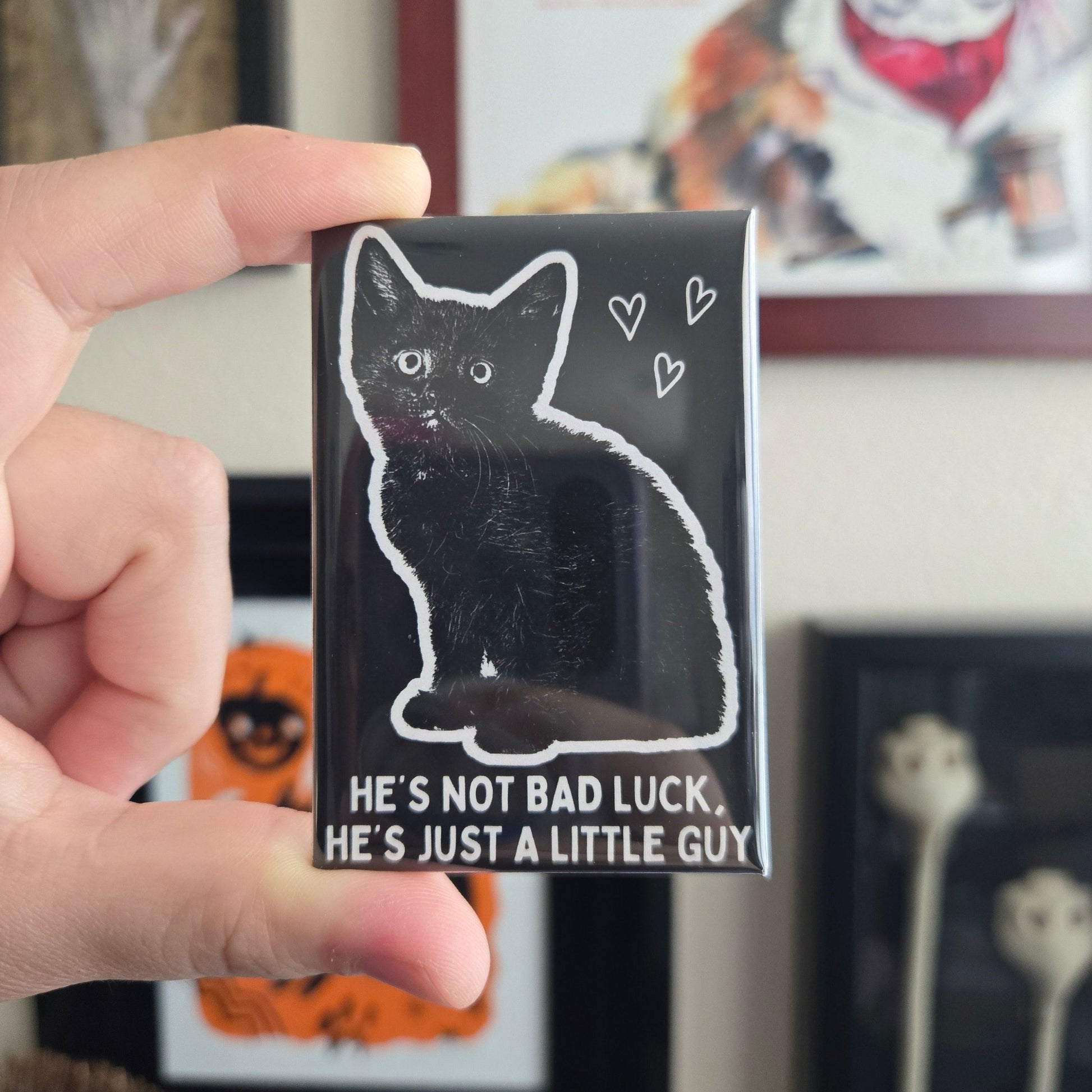 Hand holding a rectangular magnet with black and white cat illustrations reading 'He's not bad luck he's just a little guy'.
