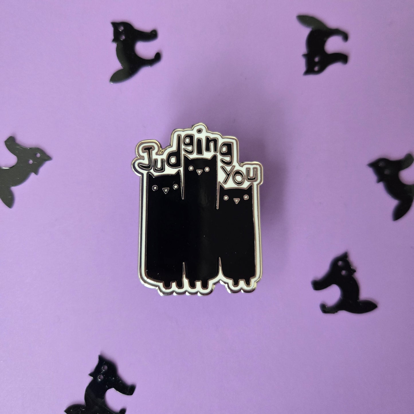 Black and white enamel pin reading Judging You with an illustration of 3 cats, displayed on a purple background with small black cat confetti.