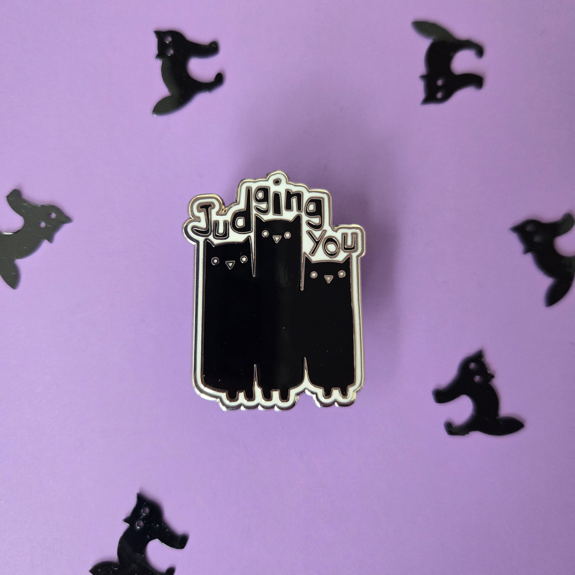 Black and white enamel pin reading Judging You with an illustration of 3 cats, displayed on a purple background with small black cat confetti.