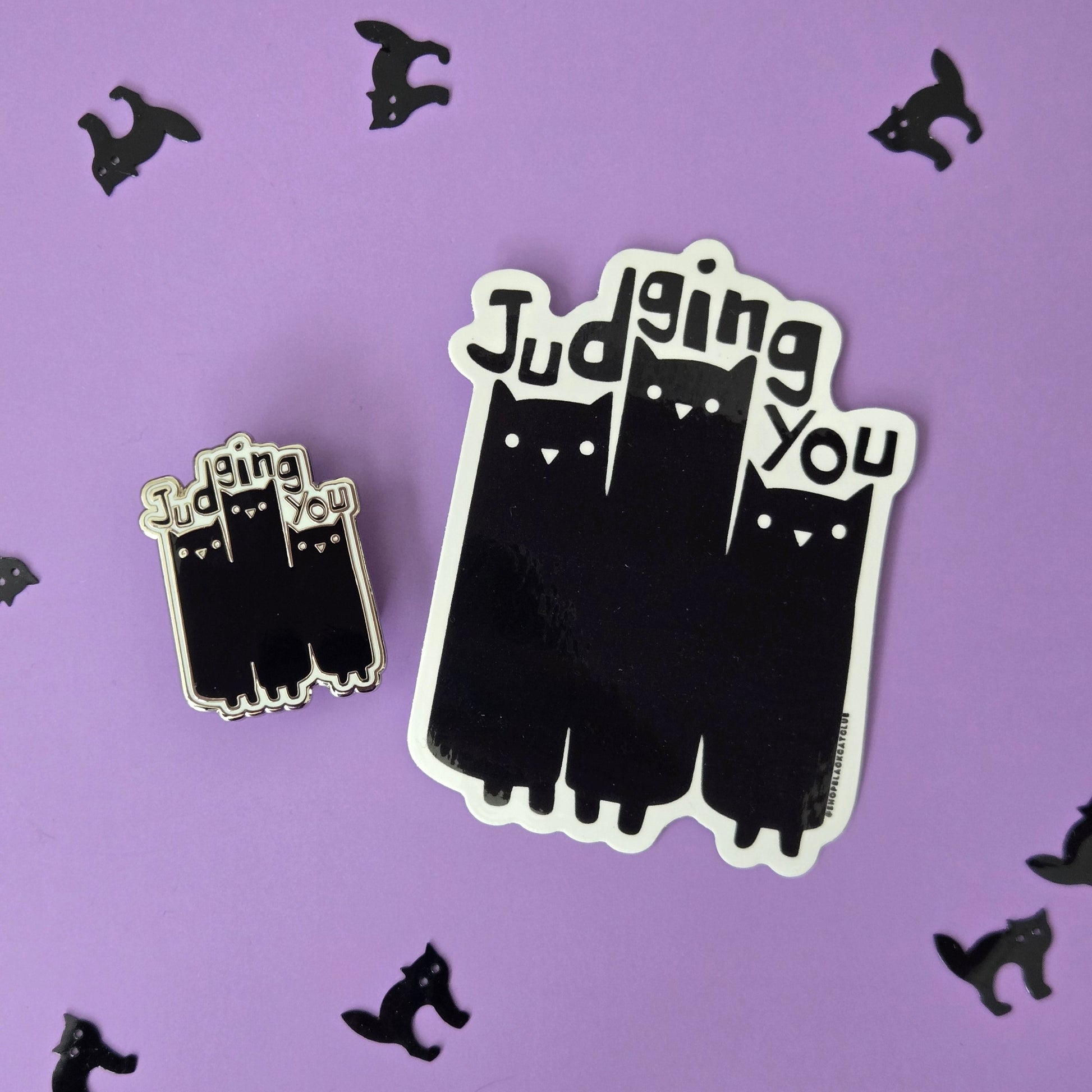 Black and white enamel pin and sticker reading Judging You with an illustration of 3 cats, displayed on a purple background with small black cat confetti.