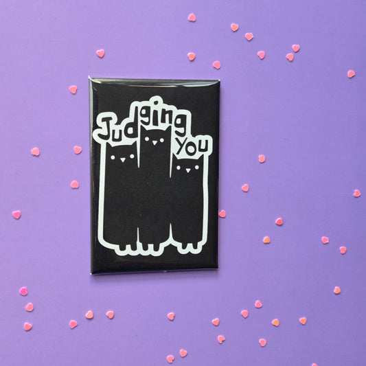 A black fridge magnet with white text and graphics showing three cats and the phrase 'Judging You' displayed on a purple background with pink sprinkles around it.