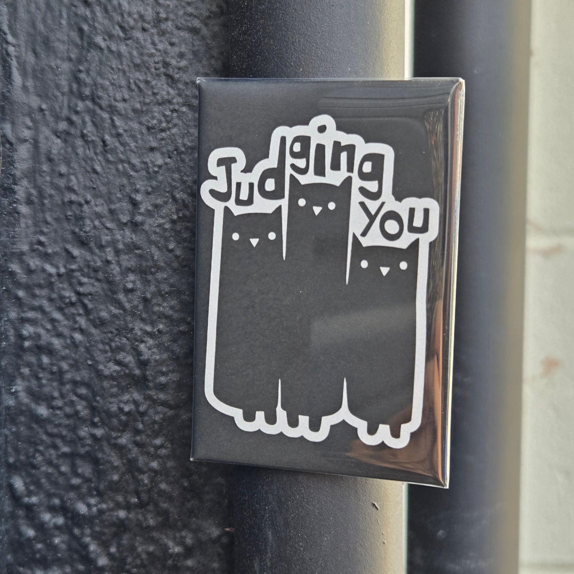 A black fridge magnet with white text and graphics showing three cats and the phrase 'Judging You'