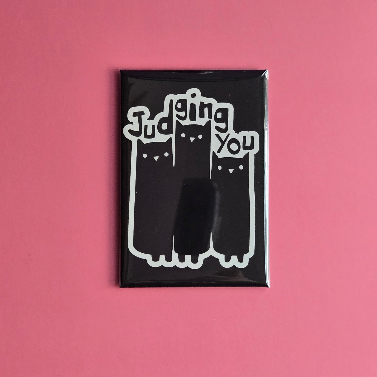 A black fridge magnet with white text and graphics showing three cats and the phrase 'Judging You'