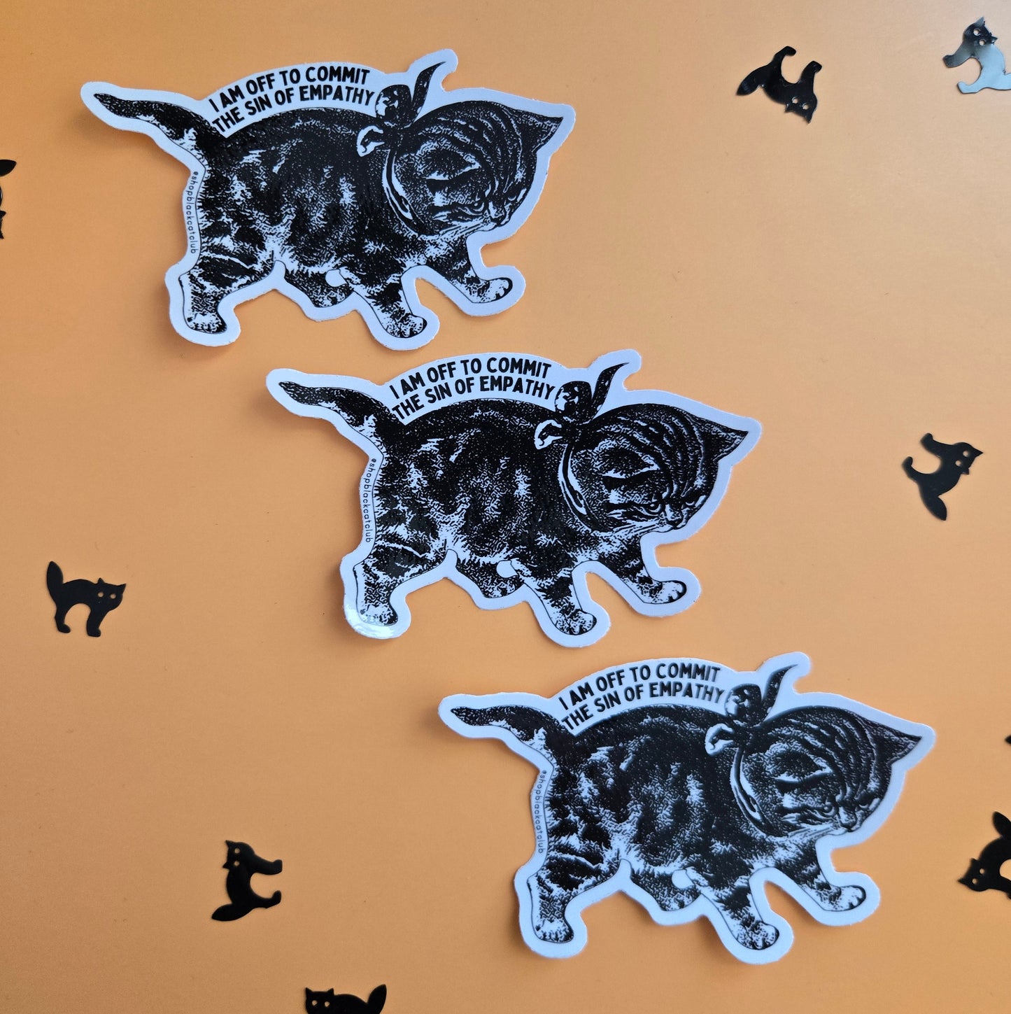 Three identical die-cut stickers featuring a black-and-white sketch of a walking cat with the text 'I AM OFF TO COMMIT THE SIN OF EMPATHY' on a warm orange background, surrounded by small black cat silhouettes.