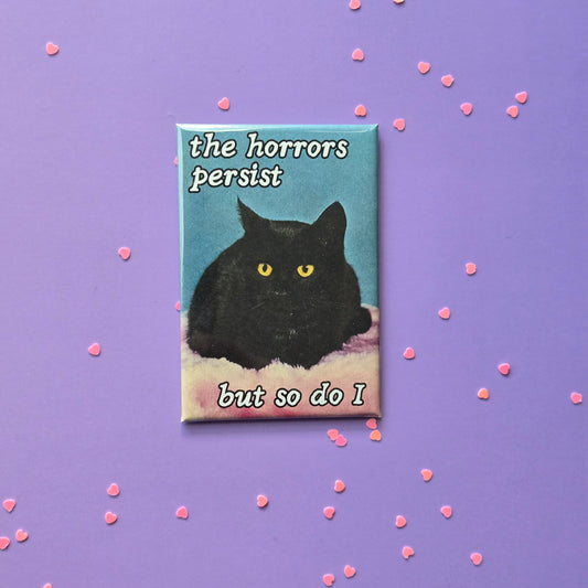 A rectangular fridge magnet featuring an photo of a black cat with the text 'the horrors persist but so do I' on a purple background with small pink heart-shaped decorations.